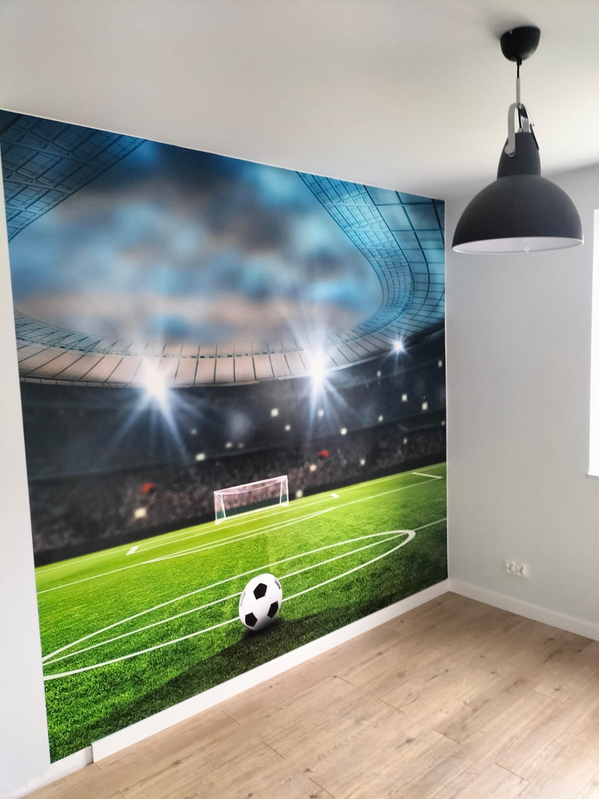 Fototapeta z motywem stadionu piłkarskiego na ścianie w pokoju, widoczna lampa sufitowa i panele podłogowe.