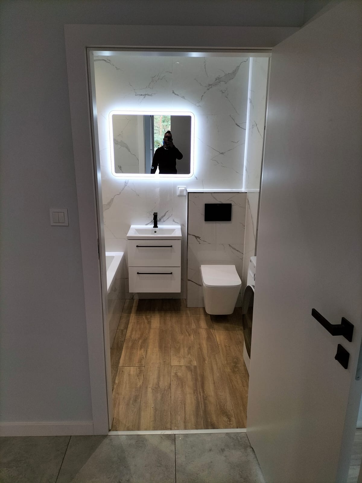 Nowoczesna łazienka po remoncie z białymi marmurkowymi płytkami, drewnopodobną podłogą, podświetlanym lustrem i wiszącą toaletą; widoczny fragment pralki i odbicie fotografa w lustrze.