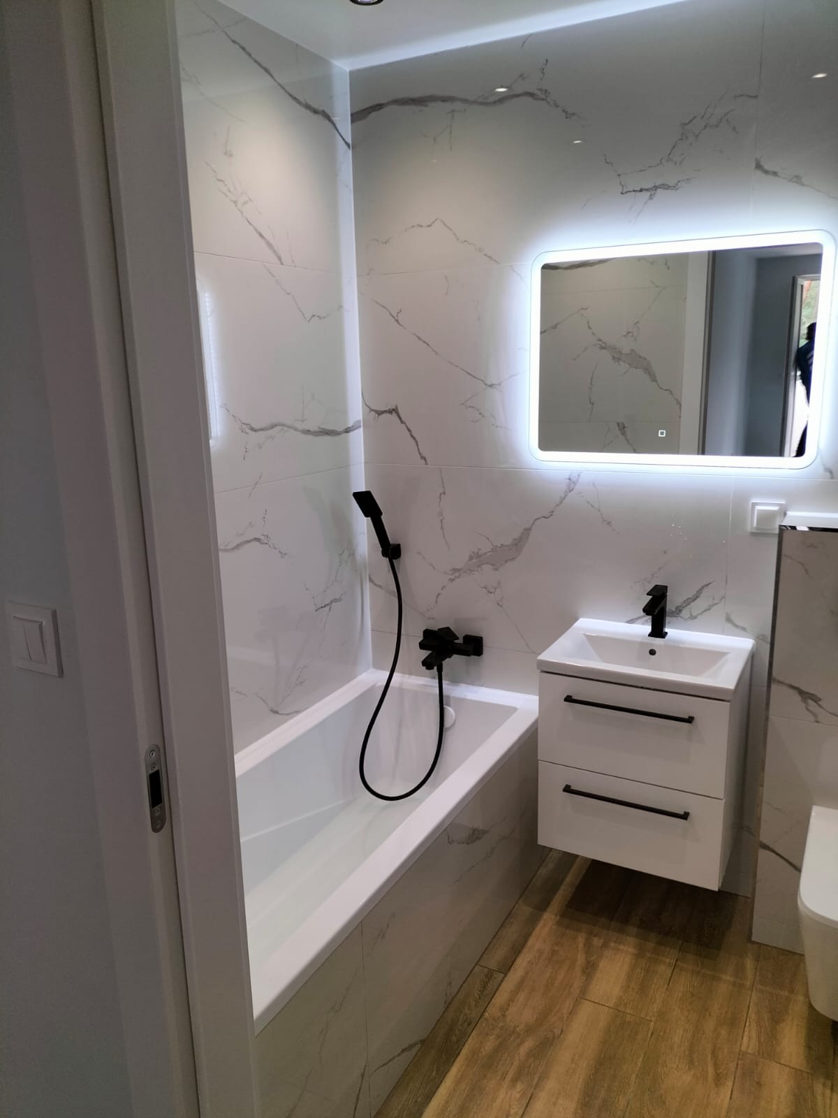 Nowoczesna łazienka z białymi płytkami imitującymi marmur, czarną armaturą i podświetlanym lustrem nad białą szafką z umywalką.