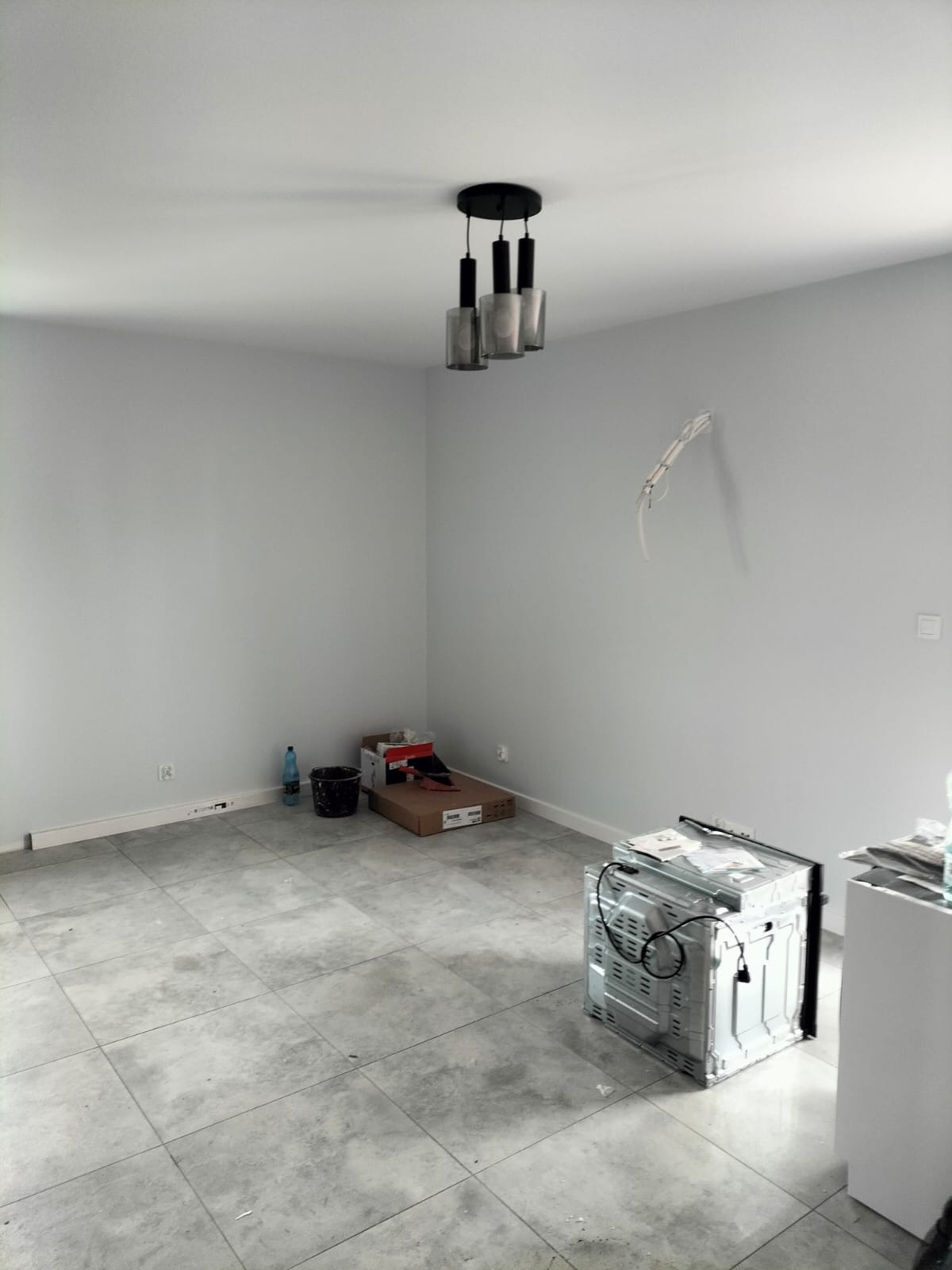 Pusty pokój z szarymi ścianami i płytkami podłogowymi, wiszącą lampą, kartonami i niedokończonymi kablami elektrycznymi, sugerujący stan przed remontem lub w trakcie prac wykończeniowych.