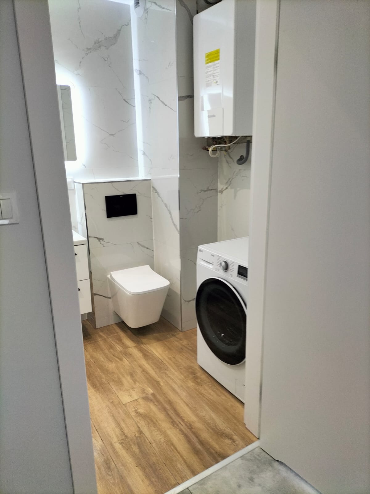 Wykończona łazienka w Lubinie z drewnopodobnymi panelami podłogowymi, wiszącą toaletą, pralką i marmurowymi płytkami ściennymi z widocznym bojlerem.