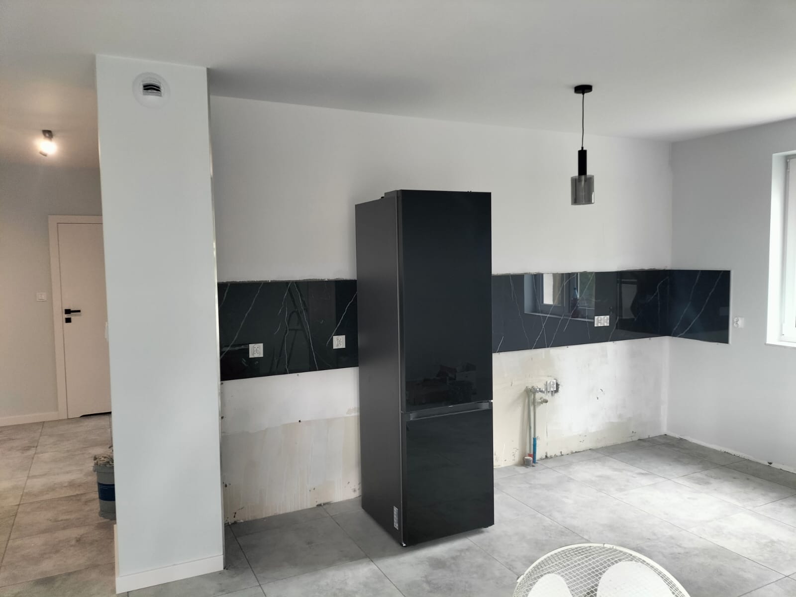 Częściowo wykończona kuchnia z czarnymi panelami imitującymi marmur nad blatem, czarną lodówką i szarymi płytkami podłogowymi, widoczne ślady po pracach remontowych na ścianach.