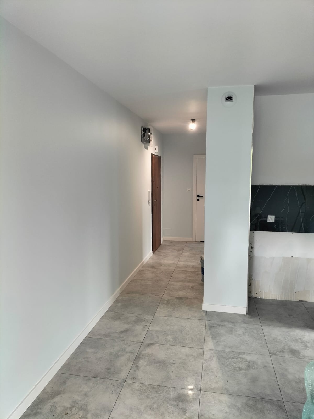 Wnętrze mieszkania po remoncie, świeżo pomalowane ściany w odcieniach szarości, ułożona szara podłoga z płytek, widoczne drzwi wewnętrzne i fragment przygotowywanej kuchni z czarnym blatem.