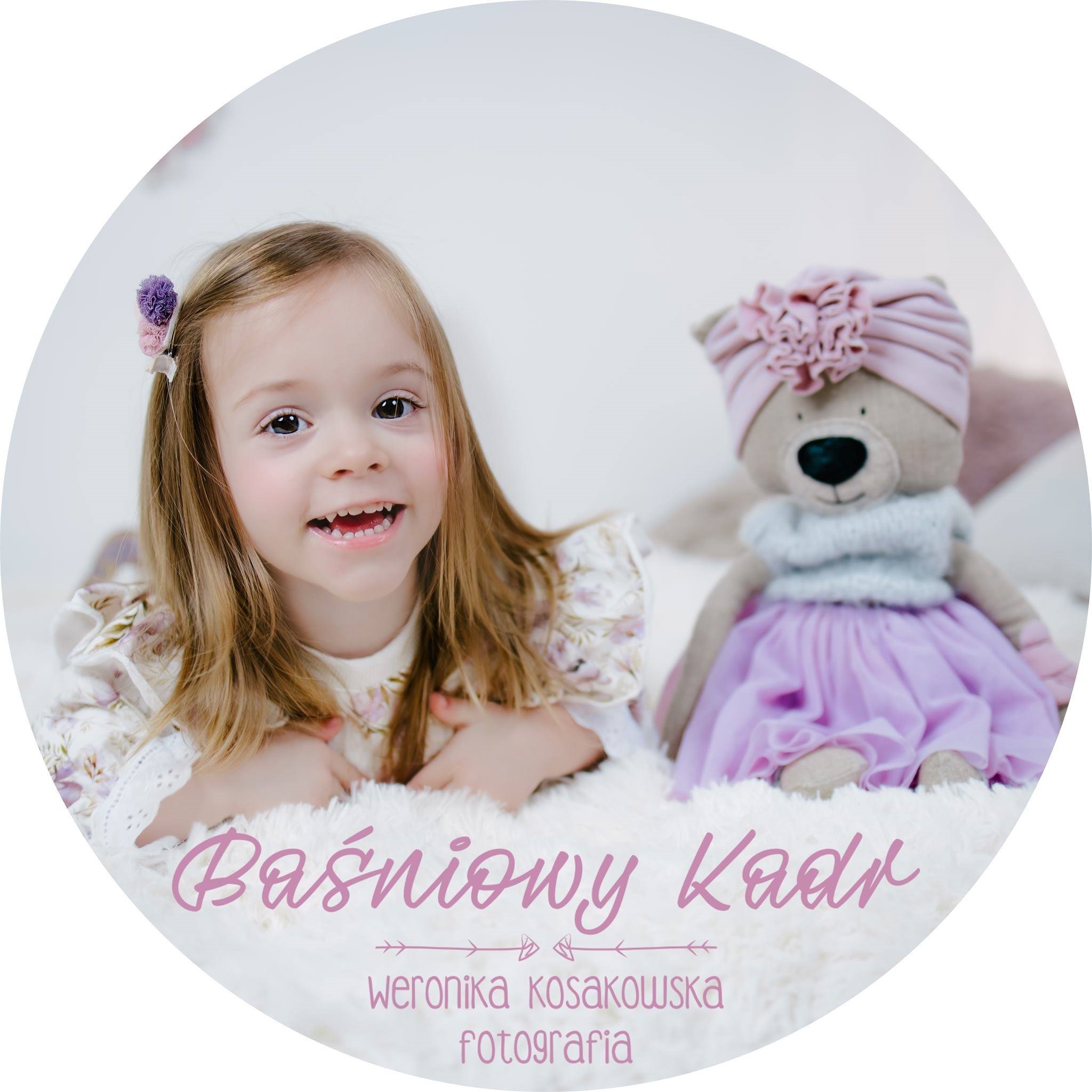 Uśmiechnięta dziewczynka z blond włosami i fioletową spinką, leżąca na puszystym białym tle obok pluszowego misia w turbanie i tiulowej spódniczce, ujęcie w okrągłej ramie.