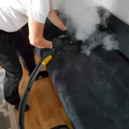 Czyszczenie parowe ciemnej sofy modułowej przez pracownika w masce i okularach, widoczna para i dysza urządzenia.