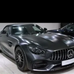 Matowy, grafitowy Mercedes-AMG GT Roadster zaparkowany w salonie, widok z przodu, z widocznym logo Mercedesa na grillu i tabliczką dealera. W tle zaparkowany inny, ciemniejszy samochód.