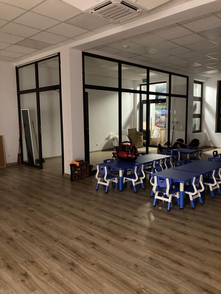 Wnętrze jasnego pomieszczenia z parkietem, niebieskimi stolikami i krzesłami dla dzieci, przeszklonymi ścianami działowymi z czarnymi ramami i klimatyzacją kasetonową w suficie.