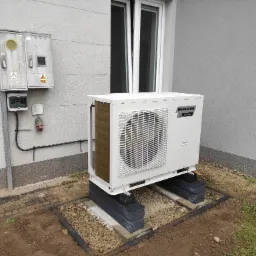 Biała jednostka zewnętrzna pompy ciepła Innova Inverter, zamontowana na betonowych bloczkach przed szarą ścianą budynku z oknem, obok skrzynki elektrycznej i instalacji.