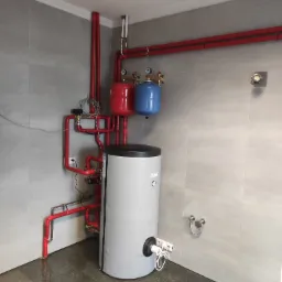 Szary bojler z czarną górą i podłączonymi przewodami elektrycznymi, obok czerwone rury instalacji grzewczej i dwa zbiorniki wyrównawcze: czerwony i niebieski, na tle szarych płytek