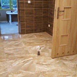 Firma Usługowo-Handlowa Jur-Dom - Wykończona łazienka z brązowymi płytkami na ścianie do połowy wysokości i beżową marmurową podłogą, widoczna otwarta drewniana ościeżnica drzwi oraz fragment pomieszczenia za nią, na podłodze stoi...