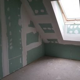 Firma Usługowo-Handlowa Jur-Dom - Poddasze z oknem dachowym w trakcie montażu płyt karton gips, widoczne szpachlowane łączenia i otwory po wkrętach, przewody elektryczne wystające ze ściany.