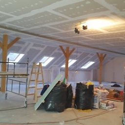 Firma Usługowo-Handlowa Jur-Dom - Poddasze w trakcie prac wykończeniowych: zamontowane płyty gipsowo-kartonowe na skośnym suficie z oknami dachowymi, widoczne drewniane słupy nośne, rusztowania i worki z materiałami budowlanymi.