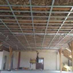 Firma Usługowo-Handlowa Jur-Dom - Stelaż sufitowy z profili metalowych, częściowo wypełniony izolacją, widoczny na poddaszu z drewnianymi belkami i oknami dachowymi, pracownik na podeście w oddali.