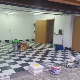 Garaż z ułożoną szachownicą z czarno-białych płytek podłogowych, widoczny odkurzacz przemysłowy, stos płytek, puszka farby, narzędzia i resztki materiałów budowlanych, otwarte drzwi garażowe.