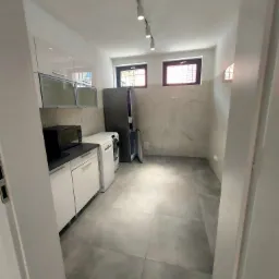 Wnętrze kuchni po remoncie, widok zza framugi drzwi: szare płytki podłogowe, białe szafki kuchenne z czarnym blatem, pralka, lodówka z otwartymi drzwiami, ściana wykończona płytami imitującymi...