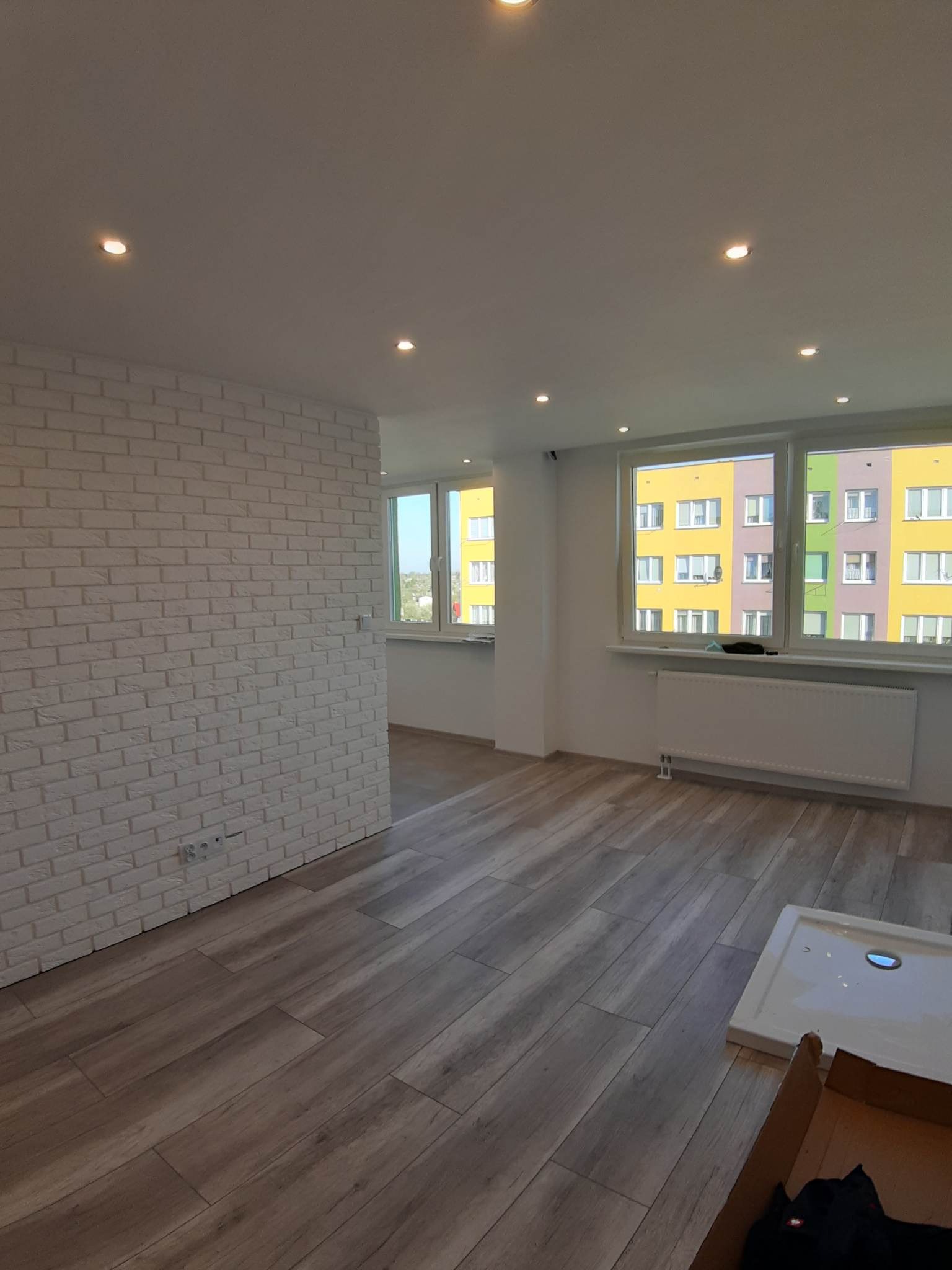 Wykończone wnętrze mieszkania z białymi cegłami na ścianie, drewnopodobną podłogą, białymi oknami z widokiem na kolorowy blok i wbudowanymi punktowymi lampami sufitowymi.