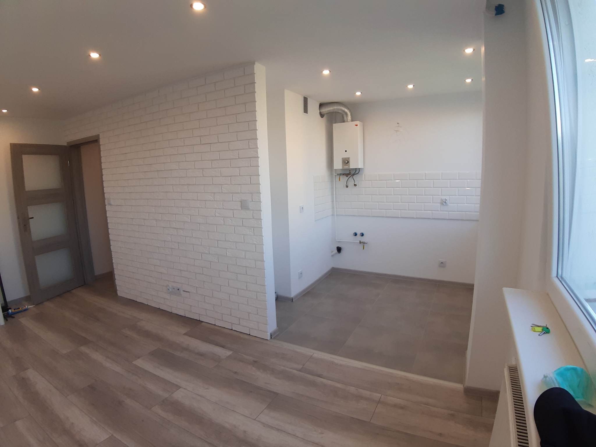 Jasne wnętrze po remoncie: ściana z białej cegły, drewnopodobna podłoga, fragment kuchni z białymi kaflami i bojlerem, klucze na parapecie.