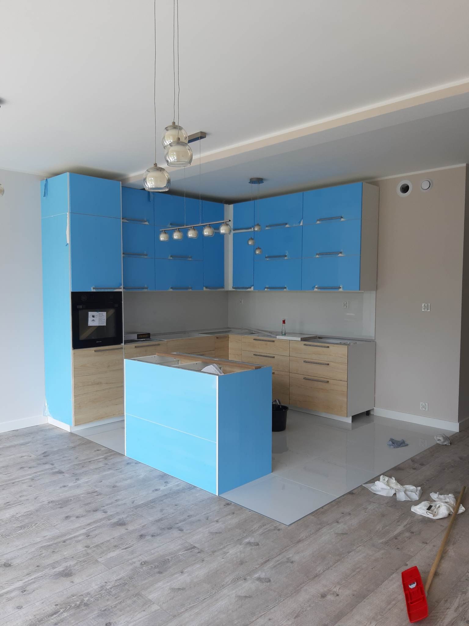 Niedokończona kuchnia z niebieskimi szafkami, drewnianymi frontami i wyspą, zabezpieczona folią ochronną, na podłodze ślady po pracach wykończeniowych, widoczny mop i szmatki.