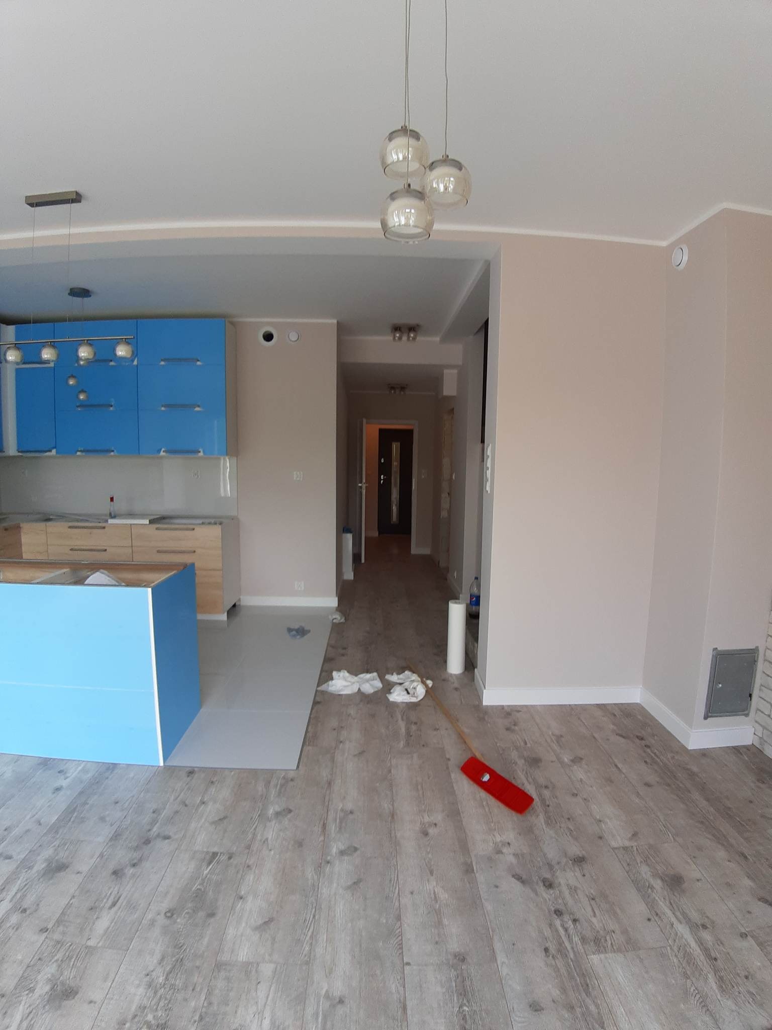 Wnętrze mieszkania w trakcie prac wykończeniowych: świeżo położona podłoga z paneli, niebieskie szafki kuchenne z błyszczącymi frontami, fragment wyspy kuchennej, ślady po remoncie w postaci plam...