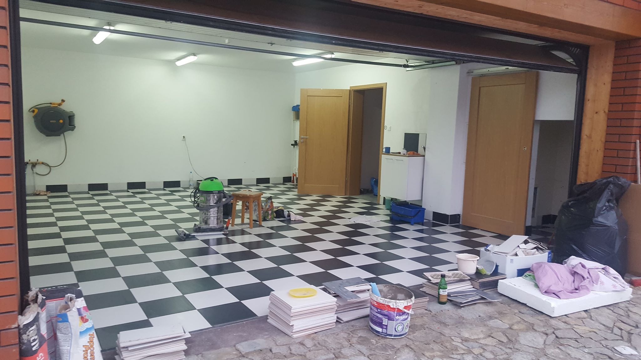 Garaż z ułożoną szachownicą z czarno-białych płytek podłogowych, widoczny odkurzacz przemysłowy, stos płytek, puszka farby, narzędzia i resztki materiałów budowlanych, otwarte drzwi garażowe.