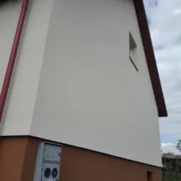 Fragment elewacji budynku z dwukolorowym wykończeniem: górna część w odcieniach bieli, dolna w brązie, widoczna skrzynka elektryczna ZG-4 oraz małe okno na górze.