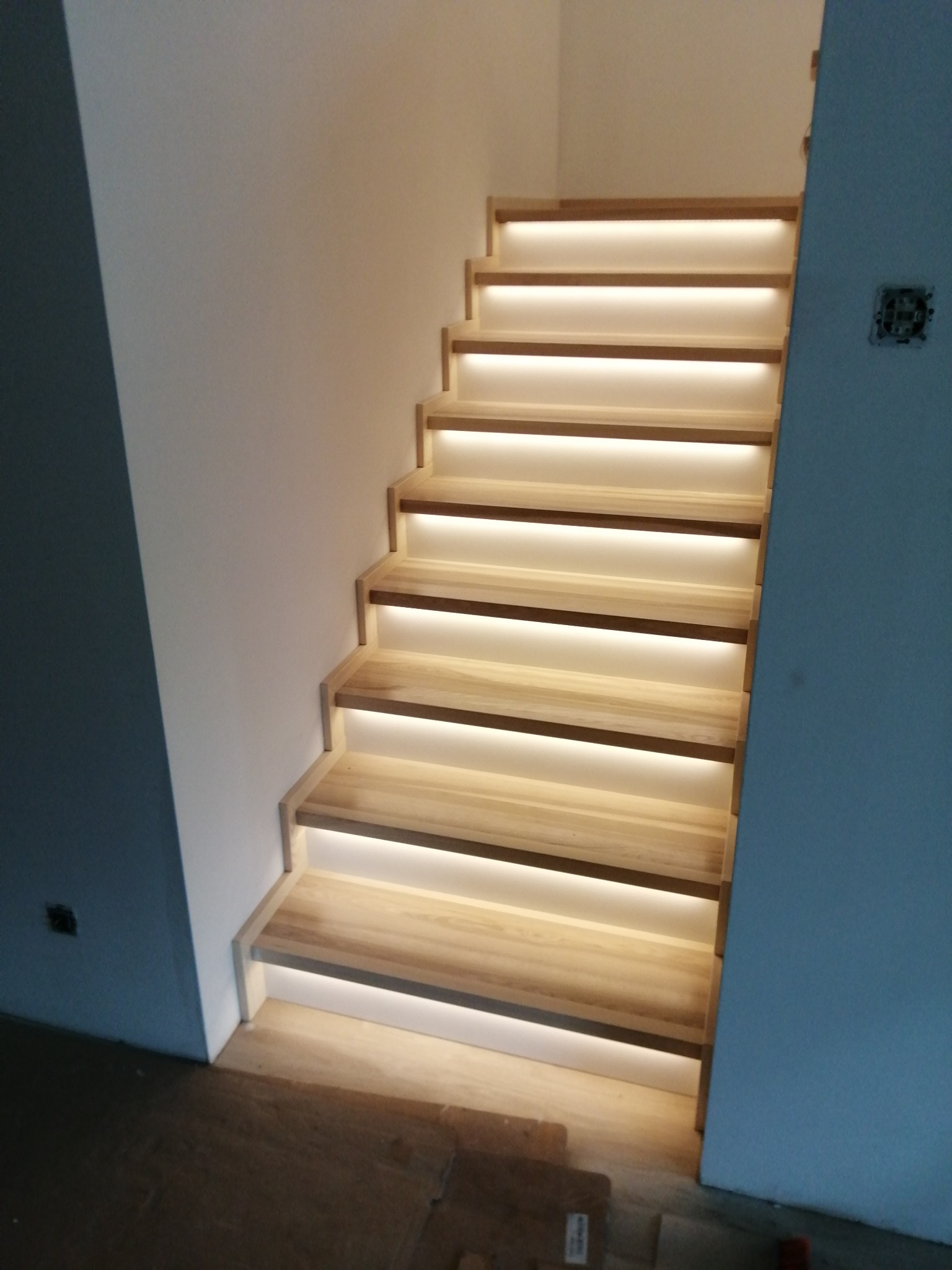 Schody jesionowe z podświetleniem LED zamontowane na betonowych schodach, widoczne niedokończone wykończenie ściany z puszką elektryczną.