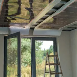 Instalacja folii grzewczej na suficie podwieszanym, widok przez otwarte okno na zewnątrz, drabina oparta o ścianę.