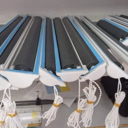 Przygotowane do montażu, zwinięte rolety z czarną tkaniną, białą obudową i niebieskimi akcentami, z łańcuszkami do regulacji, leżące na półce w magazynie.