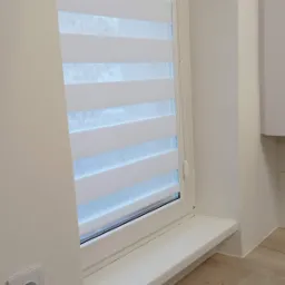 Okno z zamontowaną roletą w biało-szare pasy, widok z boku, jasne wnętrze z gniazdkiem elektrycznym na ścianie.