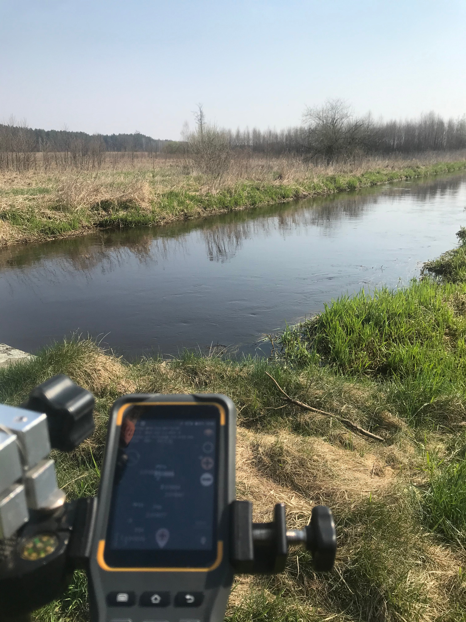 Urządzenie pomiarowe z ekranem dotykowym, ustawione na brzegu rzeki, w tle widoczny krajobraz z drzewami i łąkami.