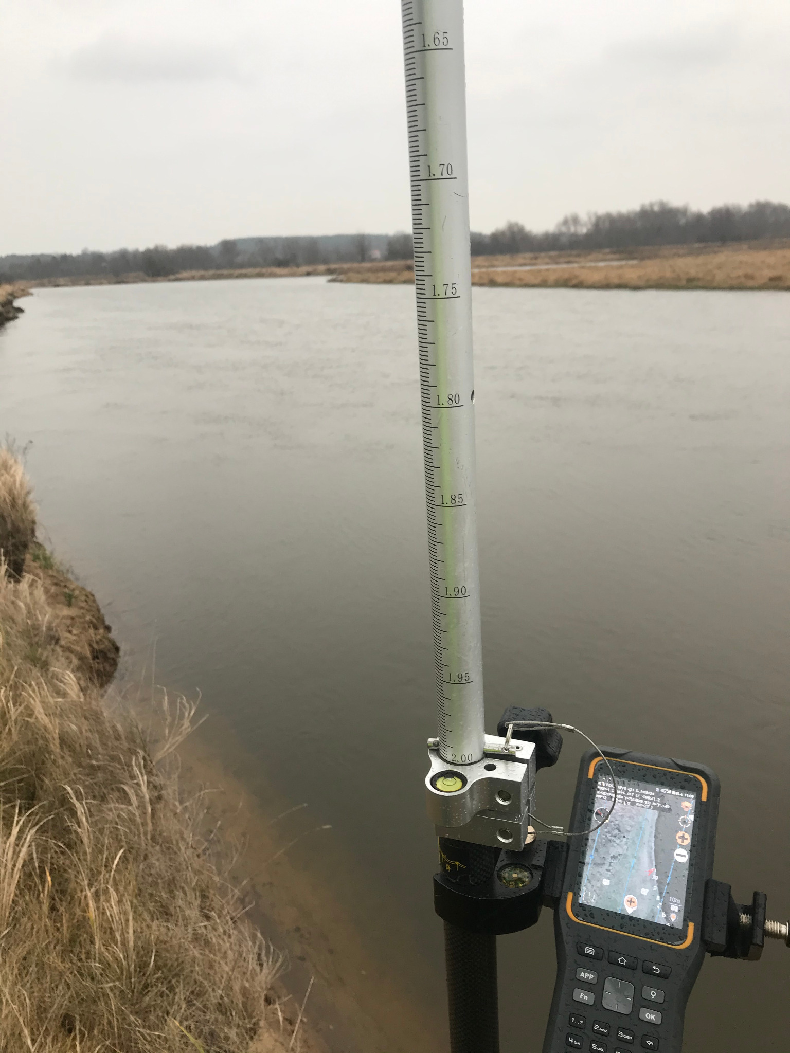 Pomiar głębokości rzeki za pomocą łaty niwelacyjnej i urządzenia GPS z mapą na ekranie, widok z brzegu porośniętego trawą, pochmurne niebo w tle.