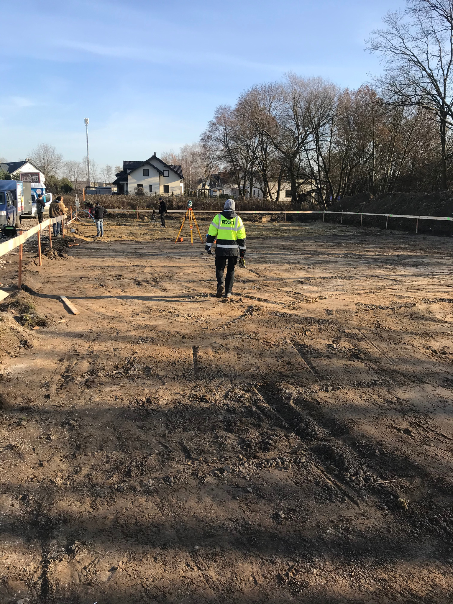 Geodeta w odblaskowej kamizelce idzie po placu budowy, w tle widoczny niwelator optyczny i ekipa budowlana.