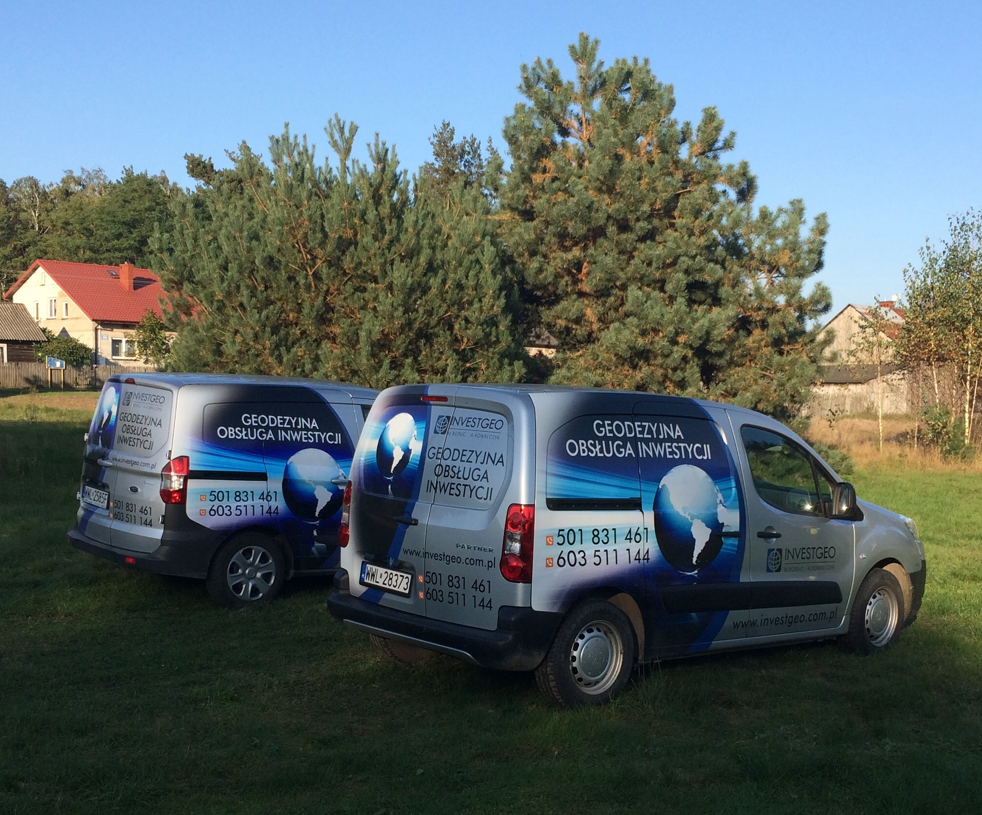 Dwa srebrne samochody dostawcze z niebieskim brandingiem 'Investgeo' i napisami 'Geodezyjna Obsługa Inwestycji' stoją na trawie w słoneczny dzień, z domami i drzewami w tle.
