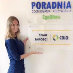 Kobieta w granatowym kombinezonie prezentuje tablicę informacyjną poradni odchudzania i odżywiania EgoSfera z logo Znaku Jakości COiO.