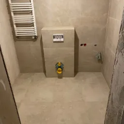 Wykończona łazienka z beżowymi płytkami imitującymi marmur, widoczny grzejnik drabinkowy, podtynkowy stelaż WC z przyciskiem spłukującym, oraz fragment ściany w stanie surowym przy drzwiach.