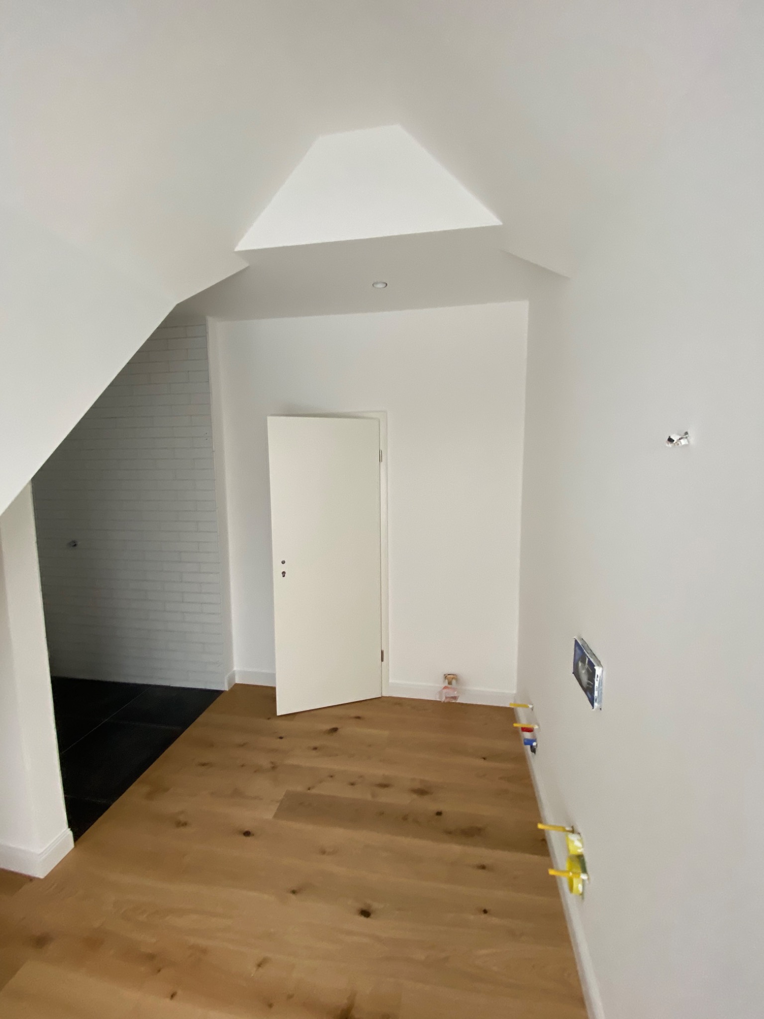 Wnętrze w trakcie wykańczania: drewniana podłoga, białe ściany z widocznymi instalacjami elektrycznymi, otwarta biała łazienka z białymi płytkami na ścianach i czarną podłogą, białe drzwi.