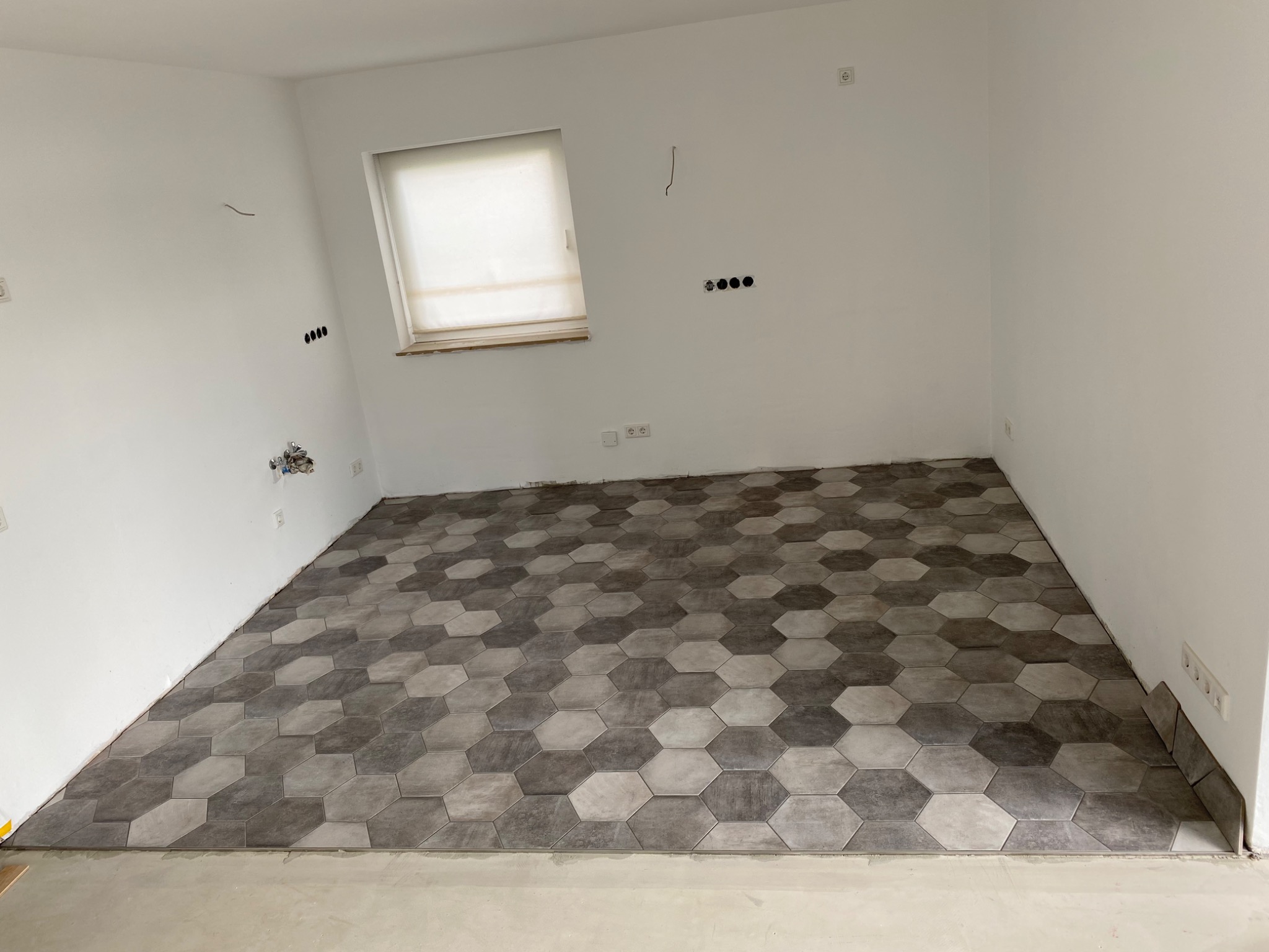 Ułożona podłoga z heksagonalnych płytek gresowych w odcieniach szarości w pustym pomieszczeniu z białymi ścianami, widoczne gniazdka elektryczne i niedokończone wykończenie przy ścianach.