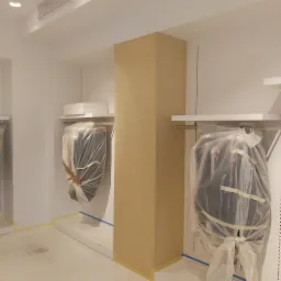 Przygotowanie do malowania biura: kolumna oklejona tapetą imitującą tkaninę, zabezpieczone folią meble na białych półkach, podłoga zaklejona taśmą malarską.