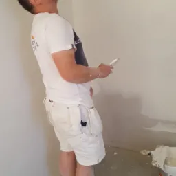 Mężczyzna w białych spodenkach i koszulce, z narzędziem w dłoni, spogląda w górę na świeżo nałożoną warstwę gładzi na ścianie, obok widoczne wiadra z materiałami i drobne zabrudzenia na podłodze.