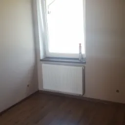 Pomalowane na beżowo ściany pokoju z ciemną podłogą, białym grzejnikiem pod oknem i widocznymi gniazdkami elektrycznymi.