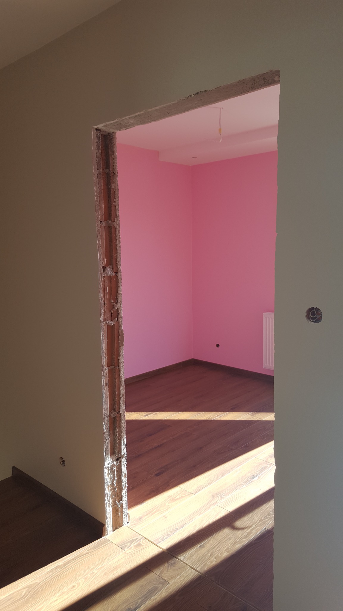 Pokój z różowymi ścianami i drewnianą podłogą widziany przez surowy otwór drzwiowy w trakcie prac wykończeniowych.