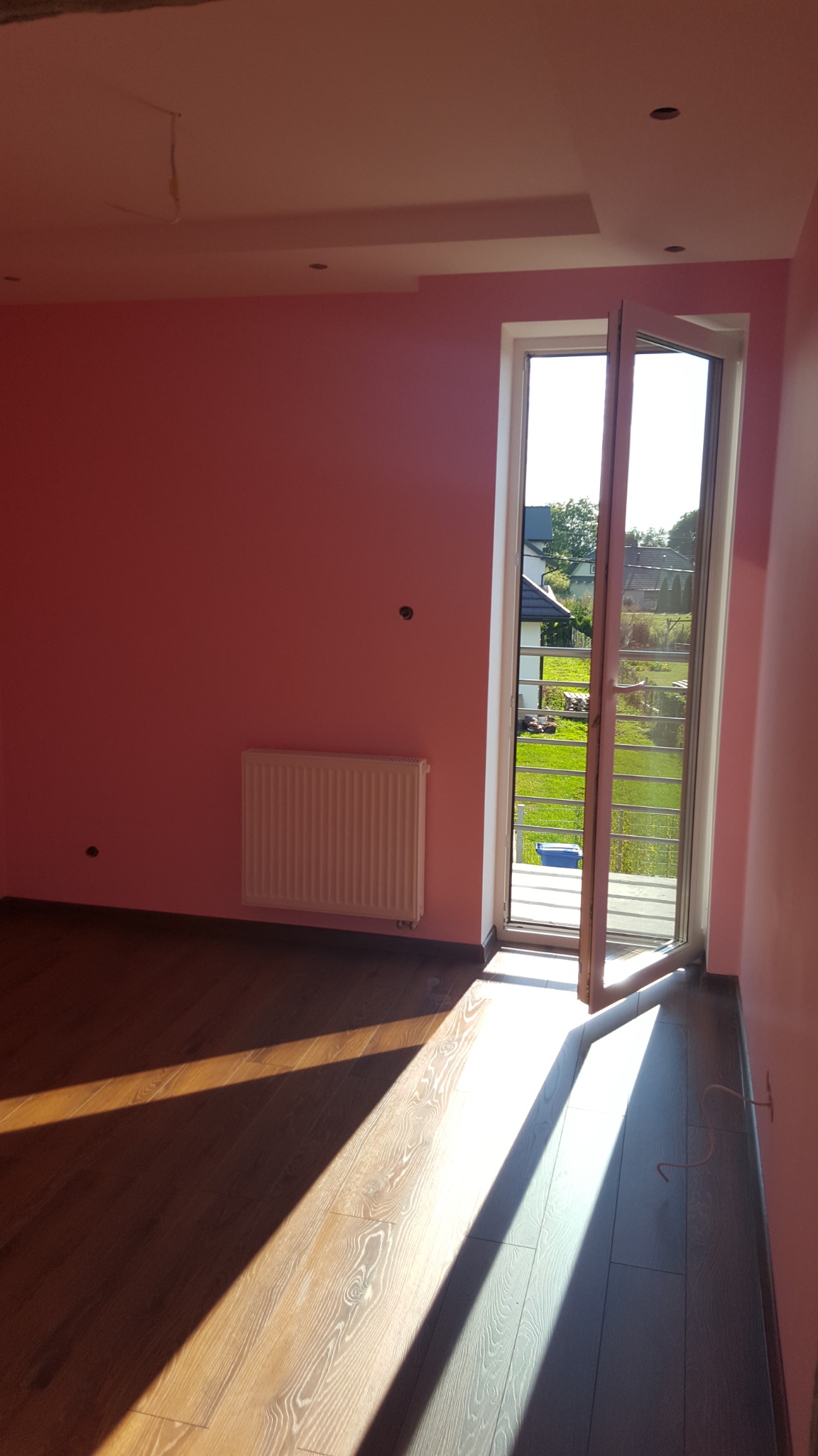 Pokój po odświeżeniu: różowe ściany, drewniana podłoga w dwóch odcieniach, biały grzejnik i otwarte drzwi balkonowe z widokiem na ogród w słoneczny dzień.