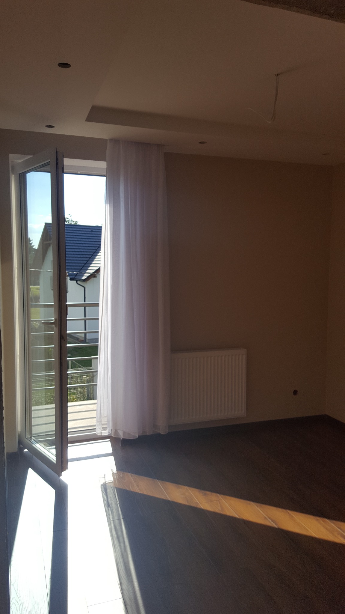 Pusty pokój z brązową podłogą, beżowymi ścianami, białym grzejnikiem i zasłoną przy przeszklonych drzwiach balkonowych, wpadające promienie słońca.