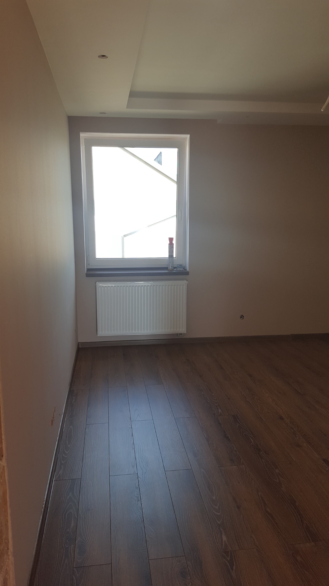 Pomalowane na szaro ściany w pustym pokoju z drewnianą podłogą, białe okno z grzejnikiem pod spodem i widocznym fragmentem sufitu z oświetleniem punktowym.
