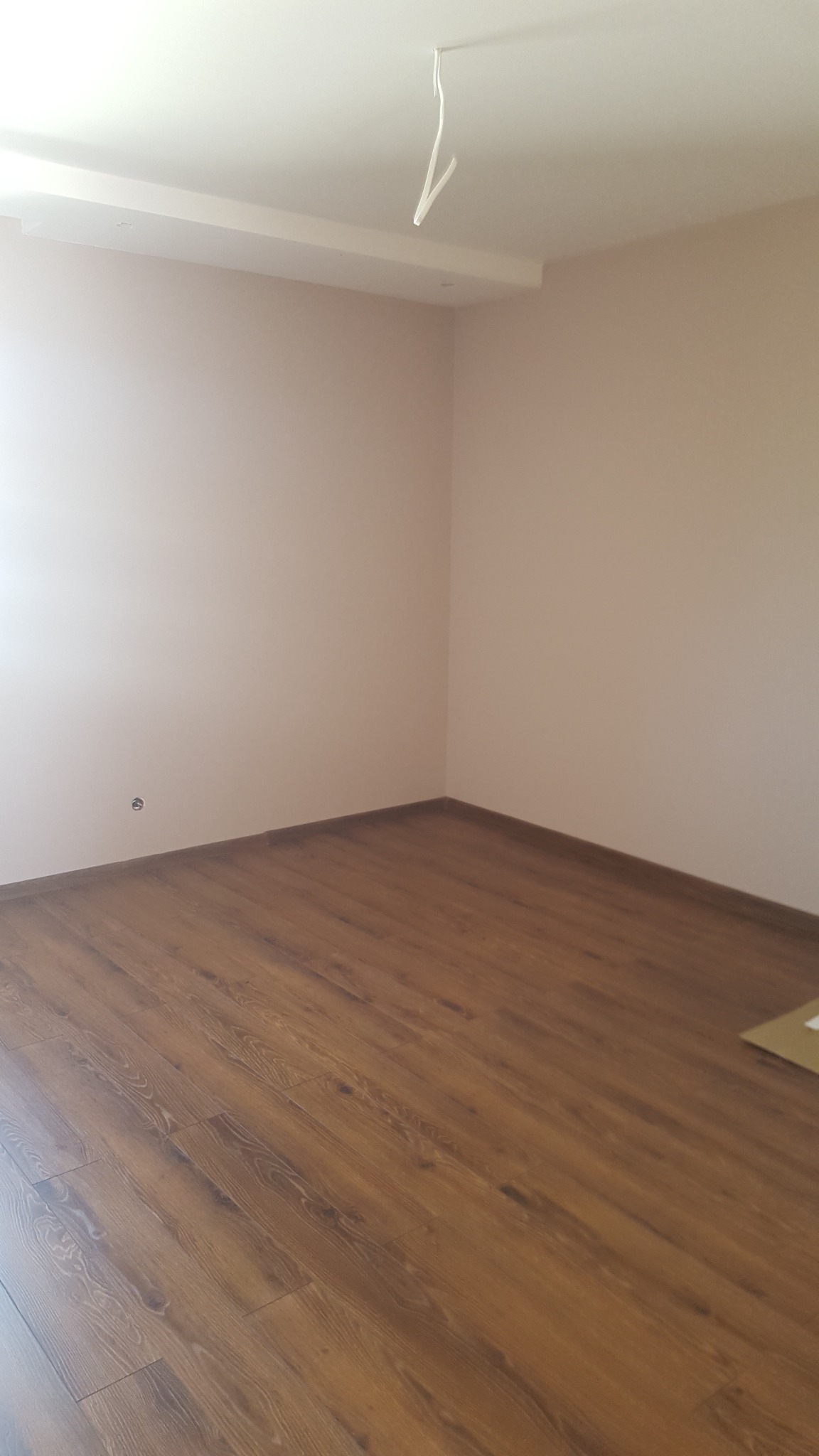 Pomalowane na jasny beż pokój z drewnianą podłogą, widoczne wystające kable z sufitu i gładka powierzchnia ścian.