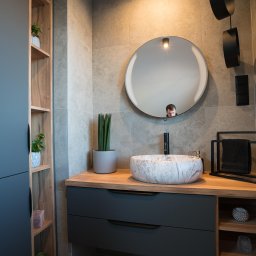 Manity Design Mateusz Łagiewa - Nowoczesna łazienka w Opolu z marmurową umywalką nablatową, czarną armaturą i okrągłym lustrem, w którym odbija się twarz osoby.