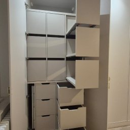 Manity Design Mateusz Łagiewa - Wnętrze szafy wnękowej z wysuwanymi szufladami i półkami, widoczne mechanizmy wysuwu, jasne kolory, fragment grzejnika, podłoga w trakcie remontu.