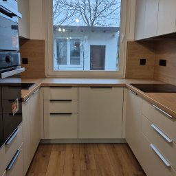 Manity Design Mateusz Łagiewa - Kompaktowa kuchnia na wymiar w jasnych kolorach z drewnianym blatem i widokiem na ogród przez okno.