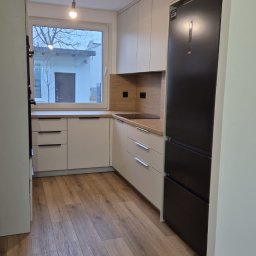 Manity Design Mateusz Łagiewa - Jasna kuchnia z drewnianym blatem, czarną lodówką i drewnianą podłogą widziana z perspektywy wejścia, z oknem wychodzącym na podwórze.