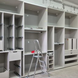 Manity Design Mateusz Łagiewa - Montaż szafy wnękowej z szufladami i półkami, widoczne prowadnice, drabina aluminiowa i narzędzia w warsztacie.